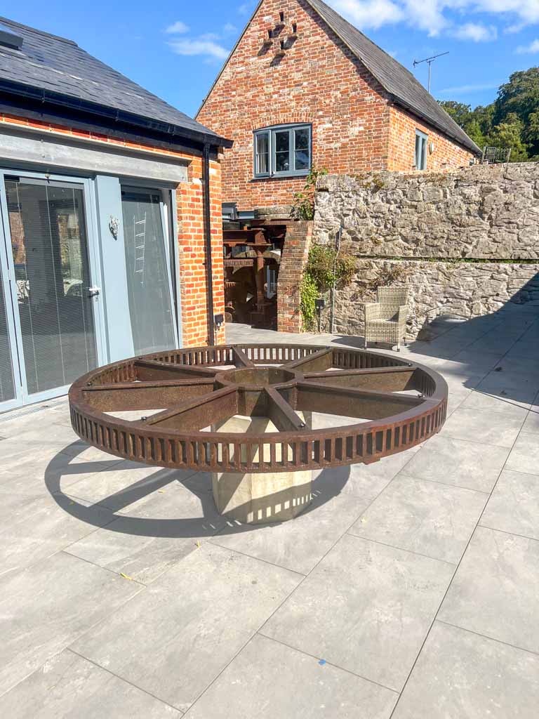 Large rusted metal gear wheel positioned on a concrete base in the centre of a sunlit outdoor patio. The gear casts a distinct shadow on pale stone paving. The patio is surrounded by red brick and stone buildings with modern windows, blending historic and contemporary architectural styles. The environment feels peaceful and carefully designed, with no visible text.